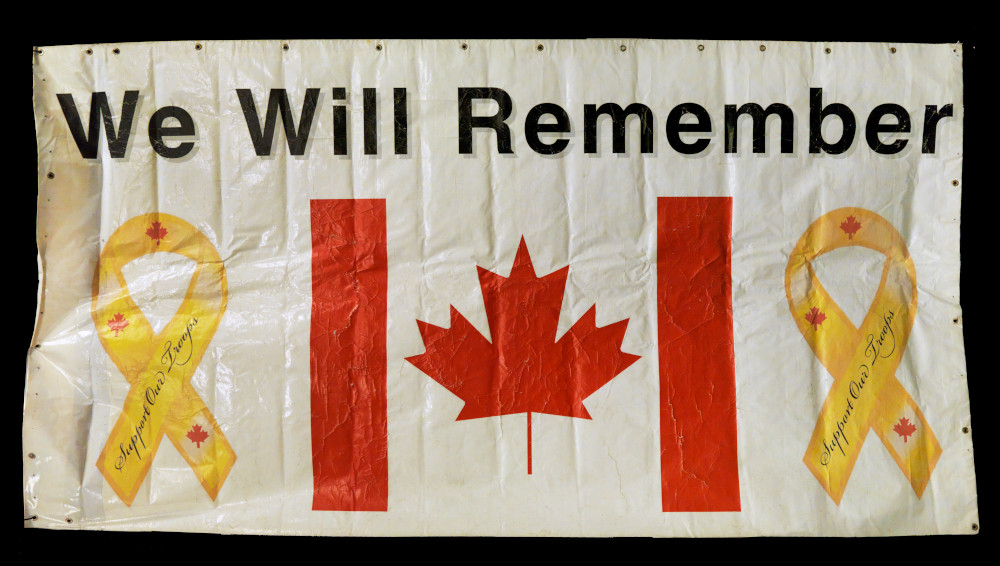 A rectangular vinyl banner with a Canadian flag flanked by two yellow ribbons reading “Support Our Troops,” beneath large text reading “We Will Remember.”